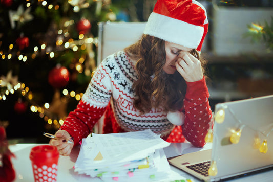 A woman in a holiday themed outfit in front of a computer and a stack of papers looks overwhelmed and puts her hand on her face