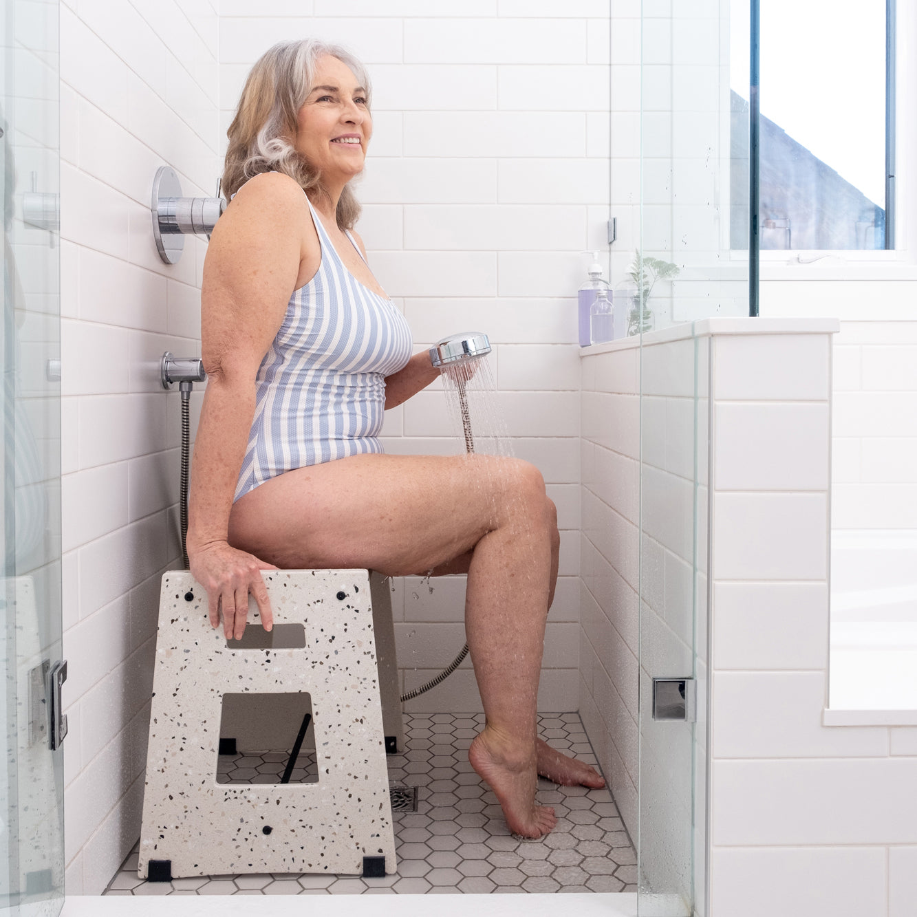 A smiling woman in a blue and white striped bathing suit sitting on the Lotic Shower Seat in terrazzo in a shower holding the detachable shower head with water running on her legs.