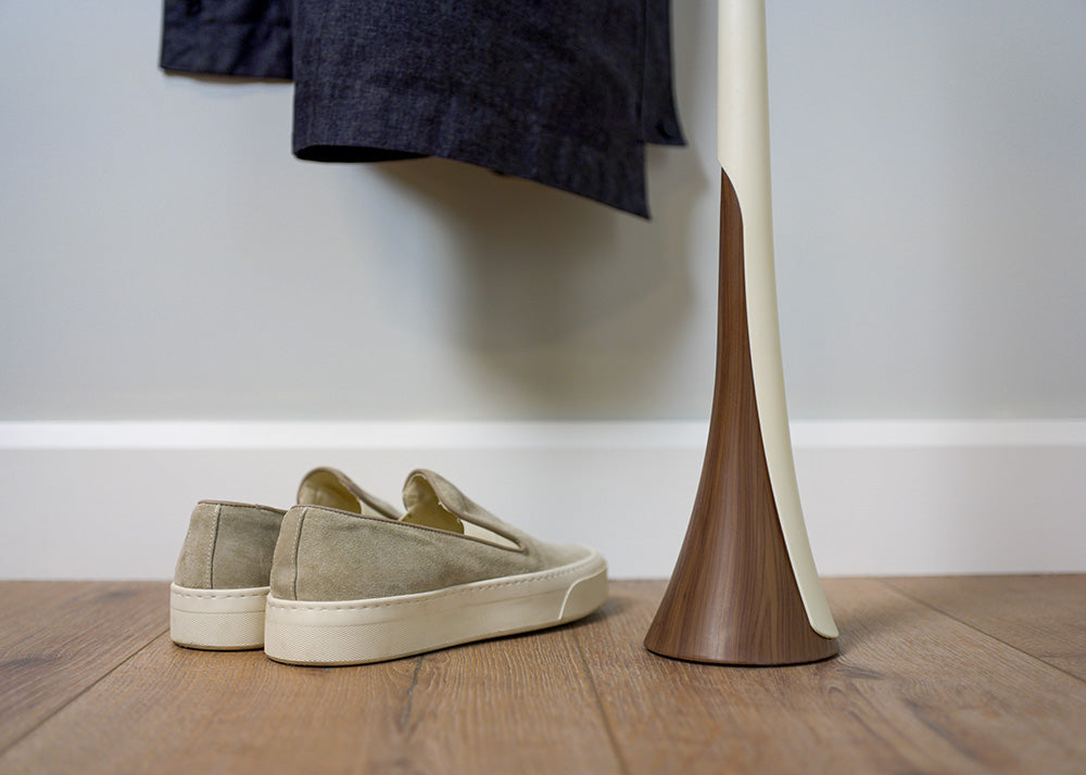 Beige slip-on shoes on a wooden floor with a standing shoehorn and a white wall and coat in the background