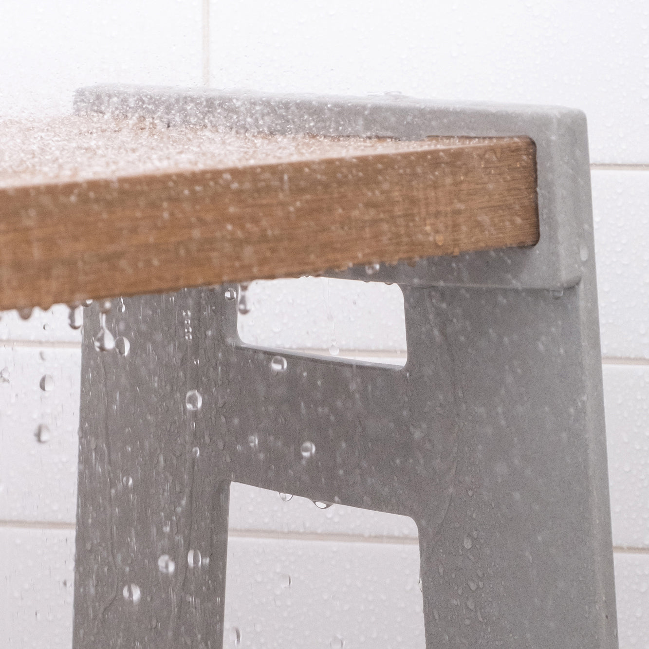 Detail of the Lotic Shower Seat in concrete grey with water falling on the grey solid surface sides and wood composite seat.