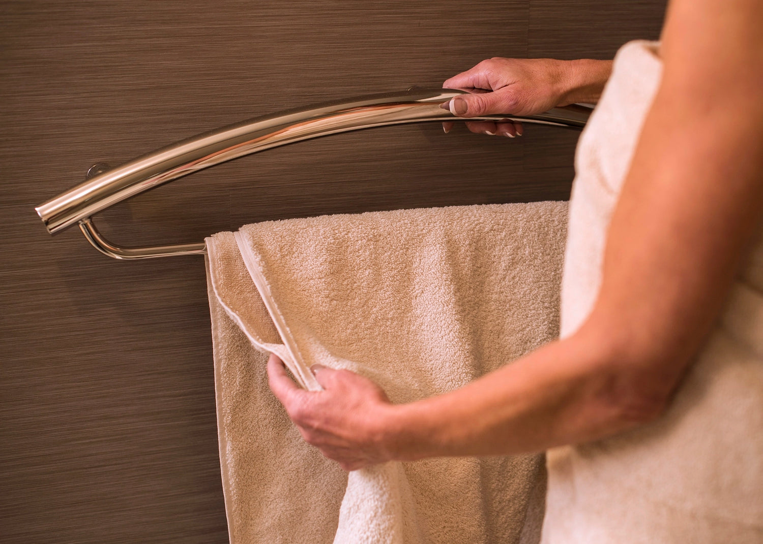 Person hanging a beige towel on a chrome towel rack attached to a chrome grab bar against a wooden wall.