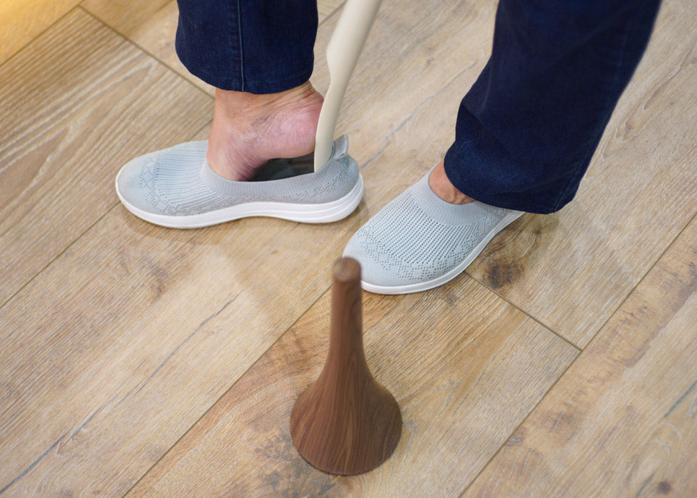 Person using a shoe horn on a wooden floor with gray slippers.